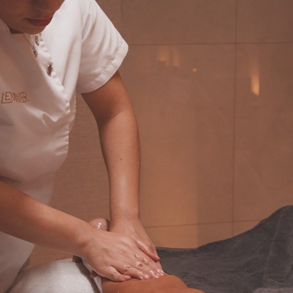 Una terapeuta de spa, vestida con uniforme blanco con el logo bordado de "LEMULIER", realiza un masaje en la pierna de una clienta utilizando aceites. La clienta está recostada sobre una camilla cubierta con toallas blancas, y su piel luce brillante por el aceite aplicado. La atmósfera es cálida y relajante, con una lámpara encendida y flores decorativas en el fondo, manteniendo la estética acogedora del entorno del spa.