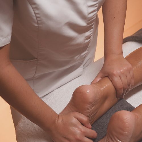 Una terapeuta de spa con uniforme blanco y el nombre "LEMULIER" bordado en el pecho realiza un masaje en el tobillo y pie de una clienta utilizando aceites esenciales. La piel luce hidratada y brillante por el producto aplicado. El entorno transmite tranquilidad, con una suave iluminación ambiental, una mesa auxiliar clara y detalles decorativos como un jarrón con flores secas que aportan un toque natural y elegante al espacio de bienestar.
