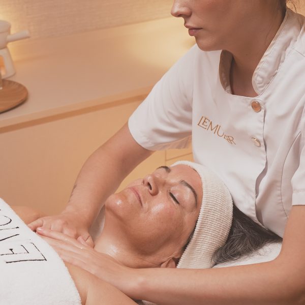 Una clienta está tumbada boca arriba en una camilla de spa, cubierta con una toalla blanca. Una terapeuta, vestida con uniforme blanco con el nombre "LEMULIER" bordado, le realiza un masaje en los hombros y la parte superior del pecho con movimientos suaves. La clienta lleva una banda en la cabeza para mantener el cabello en su lugar. El ambiente es tranquilo y acogedor, con luz cálida, una lámpara de mesa encendida y detalles decorativos como flores secas que completan la atmósfera relajante del spa.