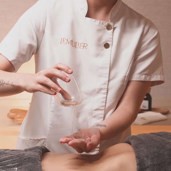 Esteticista con uniforme blanco de la marca LeMulier vertiendo aceite o loción en su mano desde un cuenco de cerámica blanca durante una sesión de masaje