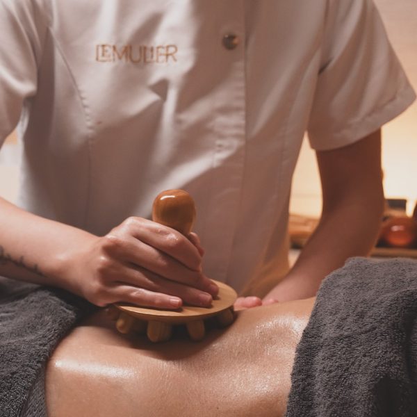 masaje utilizando una herramienta de madera con mango redondeado y protuberancias. La persona que recibe el masaje está cubierta parcialmente con una toalla oscura y su piel está aceitada, lo que indica un tratamiento relajante o terapéutico. La escena transmite una atmósfera cálida y tranquila.