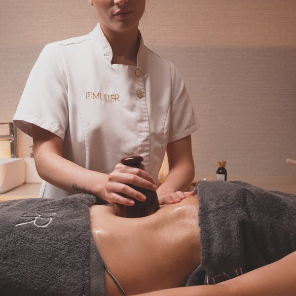 realiza un masaje utilizando una copa de madera sobre el abdomen de una persona recostada. La piel de la persona está aceitada y parcialmente cubierta con una toalla gris. El ambiente tiene una iluminación cálida y relajante, típico de un entorno de bienestar y spa.